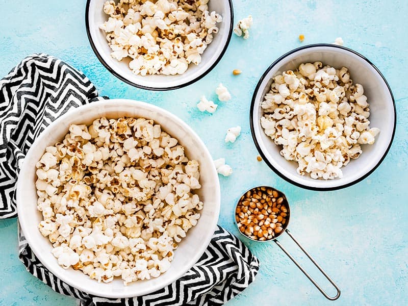 Comment faire du pop-corn moelleux et croustillant parfait avec seulement quelques ingrédients et une seule casserole. Le snack parfait, rapide et pas cher ! BudgetBytes.com Un grand bol de pop-corn avec deux bols plus petits sur le côté, avec une tasse à mesurer de grains non éclatés à côté des bols