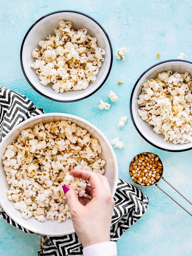 Comment faire du pop-corn moelleux et croustillant parfait avec seulement quelques ingrédients et une seule casserole. Le snack parfait, rapide et pas cher ! BudgetBytes.com Une main prenant une poignée de pop-corn dans un grand bol, deux bols plus petits sur le côté