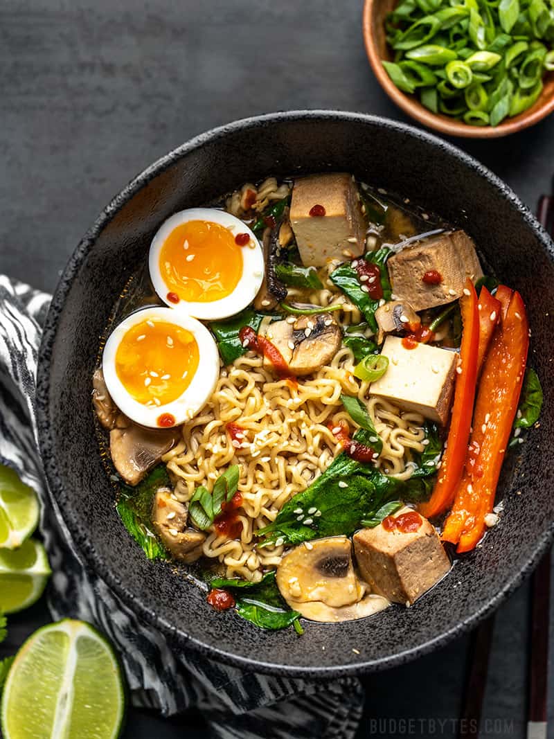 Un grand bol de ramen avec un œuf à la coque, des cubes de tofu, des épinards, du poivron et des tranches d'oignon vert en accompagnement.