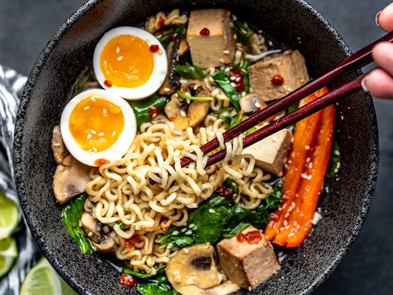 Gros plan de baguettes soulevant des nouilles d'un bol amélioré de ramen instantané, avec un œuf à la coque.