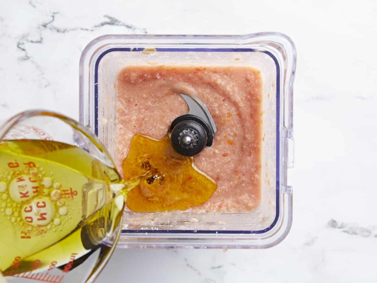 Plan aérien d'huile d'olive versée dans un bol mélangeur rempli de purée de tomates et de pain.