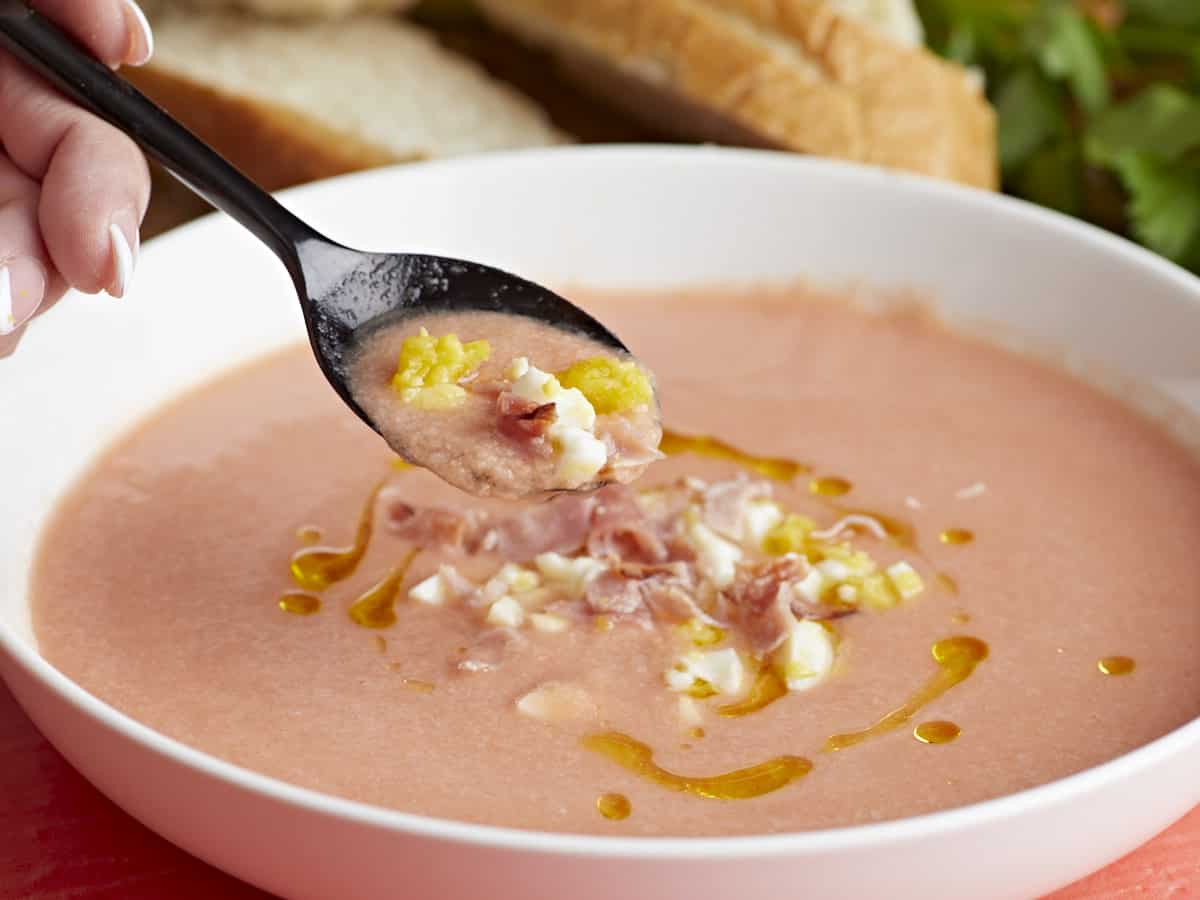 Plan latéral d'un bol blanc de Salmorejo garni d'un filet d'huile d'olive, d'œufs durs hachés et de jambon croustillant s'émiette avec une cuillère noire qui en sort.