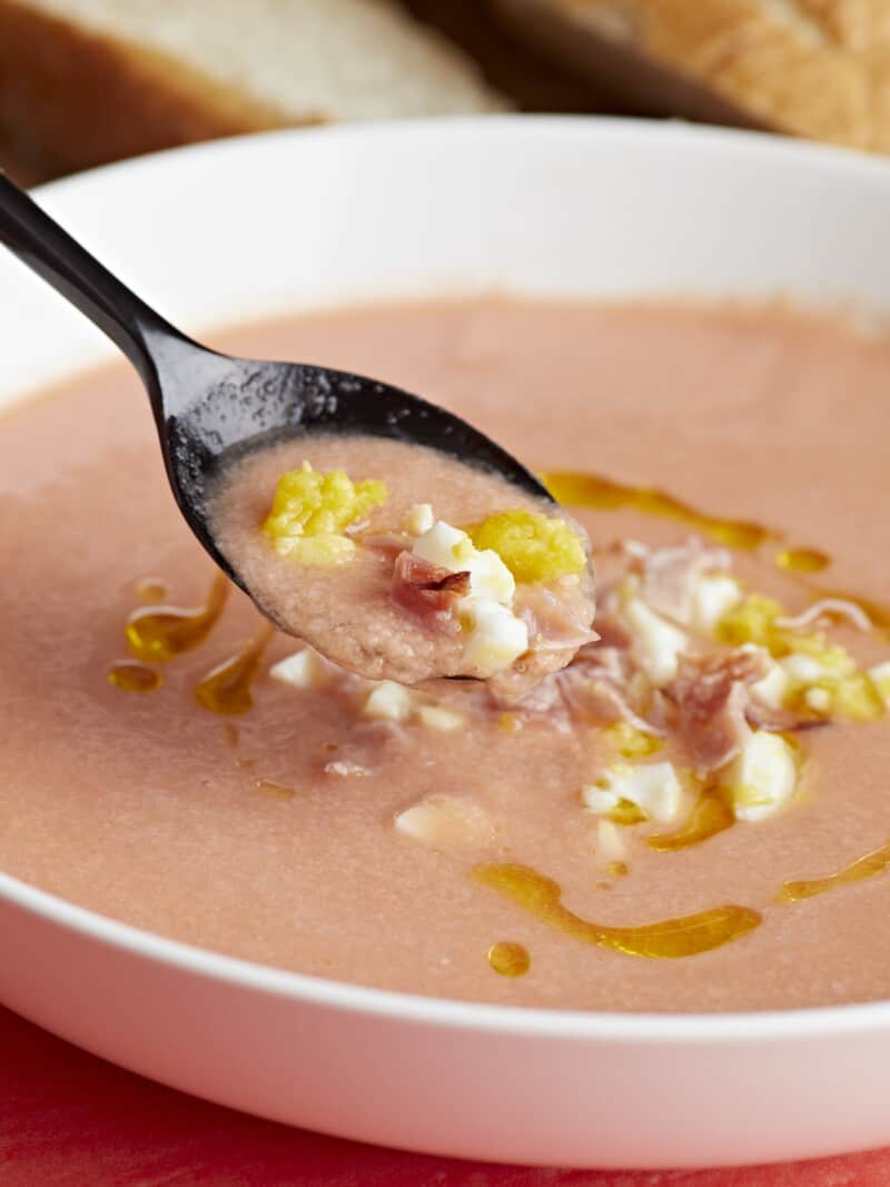Plan latéral d'un bol blanc de Salmorejo garni d'un filet d'huile d'olive, d'œufs durs hachés et de jambon croustillant s'émiette avec une cuillère noire qui en sort.
