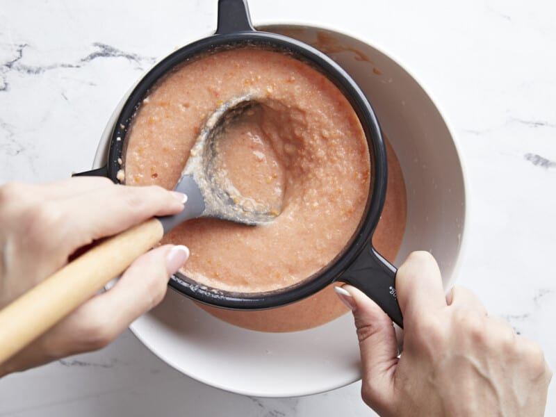 Plan aérien d'une main utilisant une louche grise pour pousser Salmorejo à travers un tamis à mailles fines avec une poignée noire dans un bol blanc.
