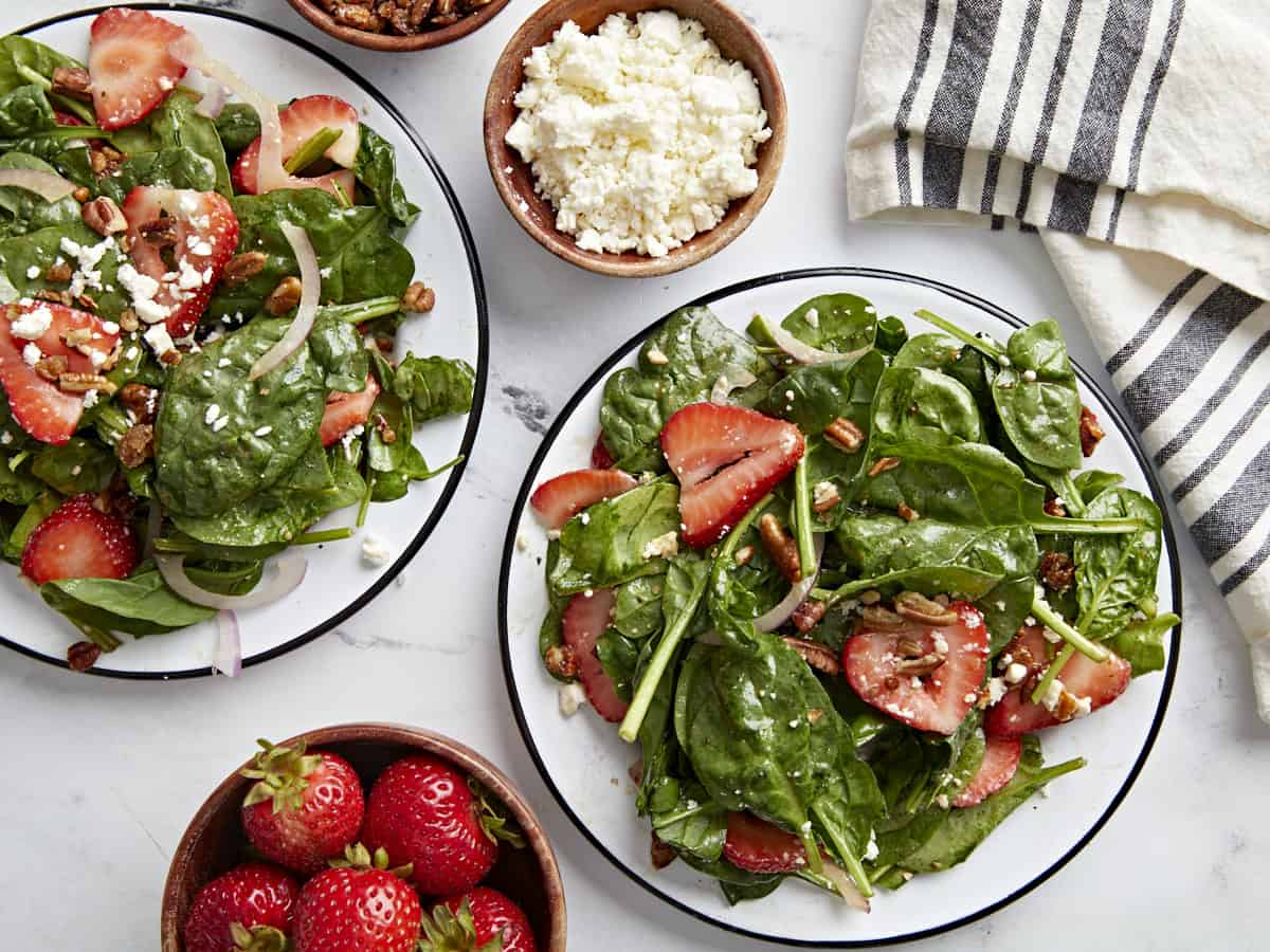 Plan aérien de deux assiettes de salade d'épinards aux fraises avec un petit bol de fraises et de fromage feta sur le côté.