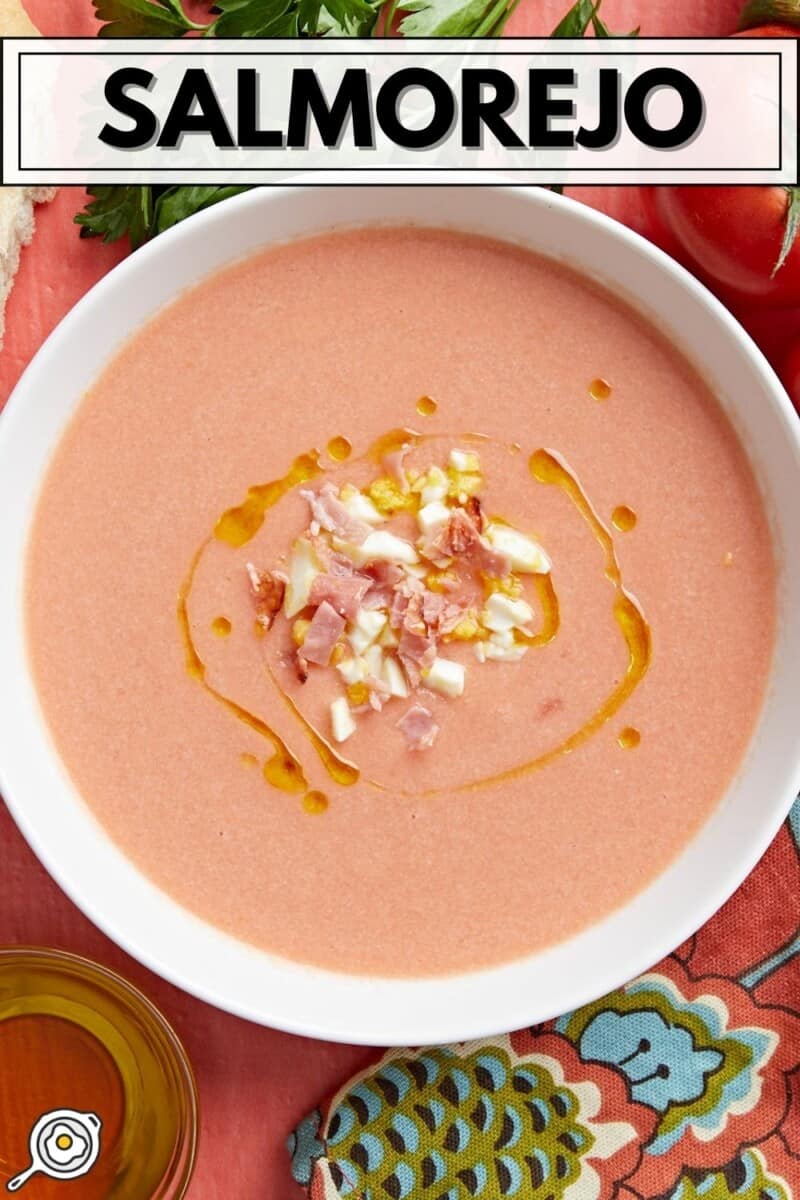 Plan aérien d'un bol blanc de Salmorejo garni d'un filet d'huile d'olive, d'œufs durs hachés et de jambon croustillant émietté.