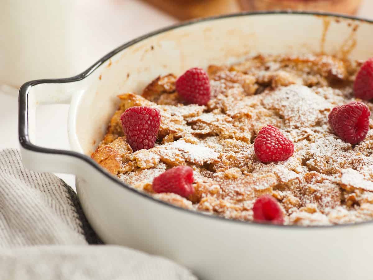 Une image horizontale en gros plan montrant une vue latérale d'un pouding au pain cuit dans un plat de cuisson rond en émail blanc garni de sucre en poudre et de framboises fraîches.