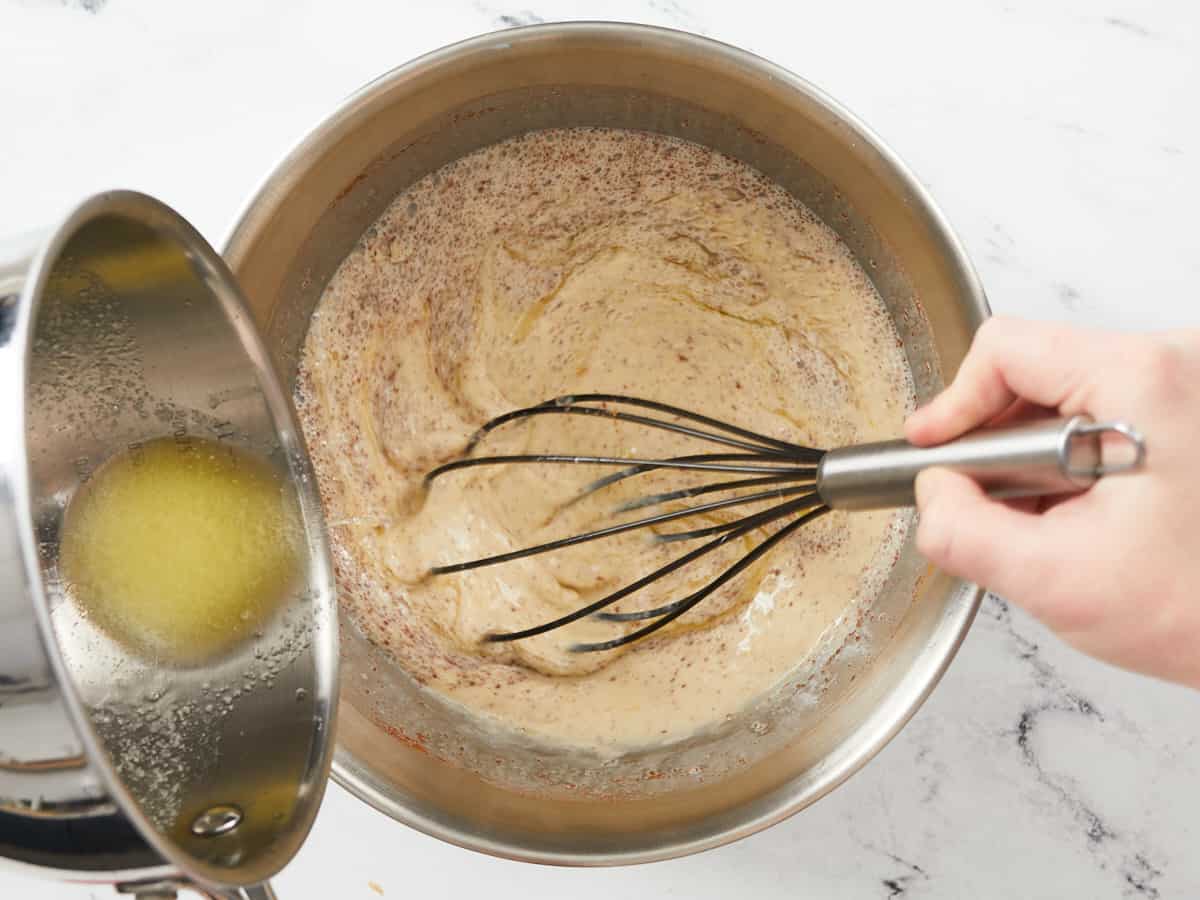 Un grand bol à mélanger en métal rempli des ingrédients de la crème au pouding au pain est fouetté tandis qu'une main hors du cadre verse du beurre fondu dans une petite casserole.