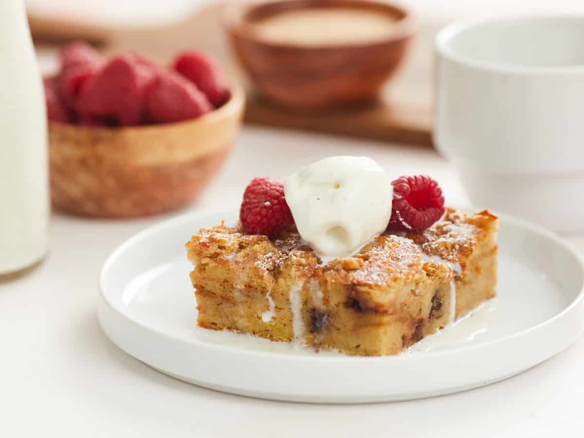 Image horizontale d'une seule portion de pouding au pain sur une assiette plate blanche garnie de deux framboises et d'une boule de glace à la vanille. En arrière-plan, il y a des bols flous d'ingrédients de garniture dans la recette, comme une petite bouteille de crème épaisse, un bol de framboises fraîches et un bol de garniture au sucre à la cannelle.