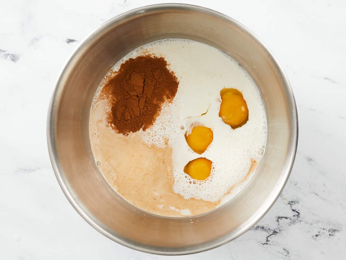 Un grand bol à mélanger en métal rempli d'ingrédients séparés pour le pudding au pain, à savoir des œufs, moitié-moitié, de l'extrait de vanille, ⅔ tasse de sucre, 2 cuillères à café de cannelle et du sel, posés sur un fond de marbre blanc.