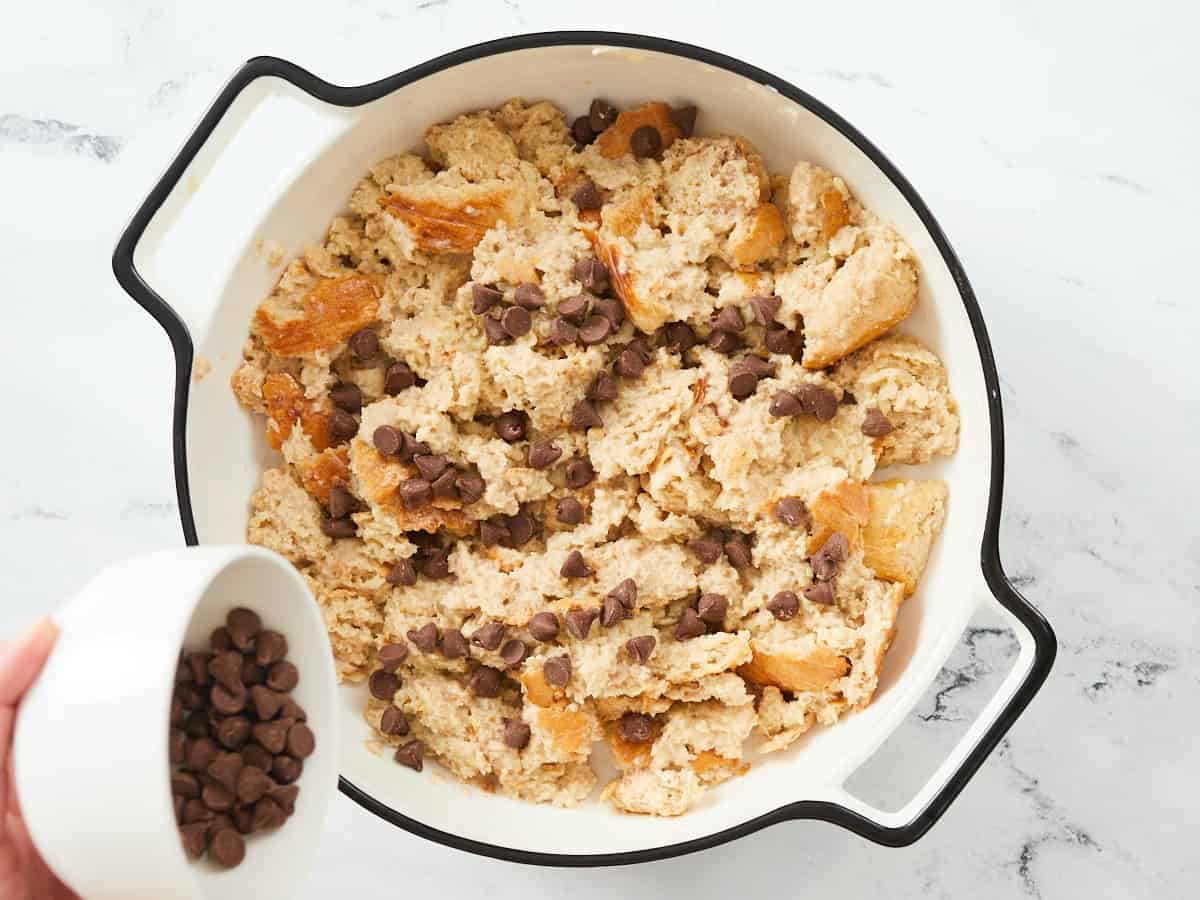 Un plat de cuisson rond en émail blanc avec une bordure noire posé sur un fond en marbre blanc. Il y a une seule couche de pain trempé et une main saupoudre des pépites de chocolat en une couche uniforme dans un petit bol blanc.