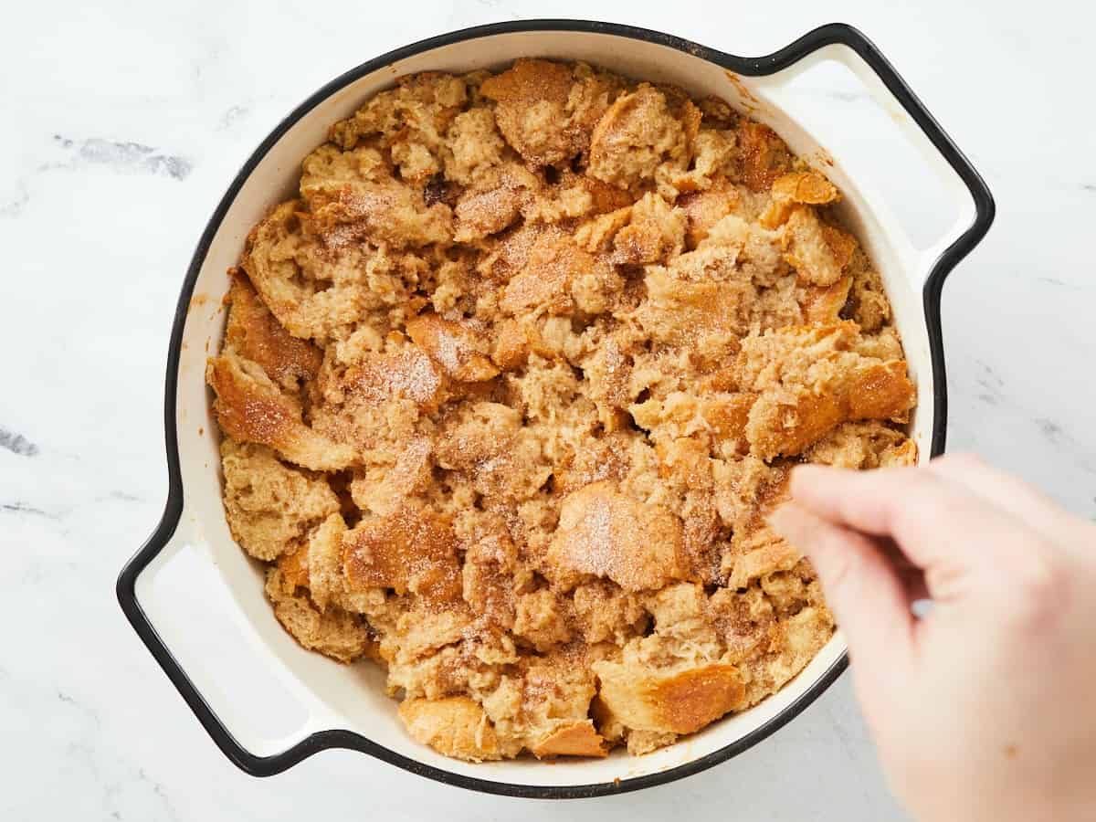 Un plat de cuisson rond en émail blanc sur fond de marbre blanc à moitié cuit. Une main dans le coin inférieur droit du cadre saupoudre le dessus de la cocotte de sucre à la cannelle.