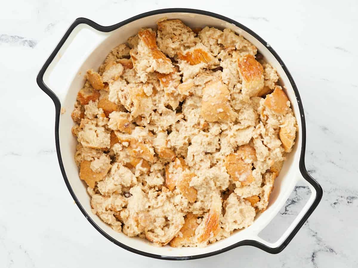 Un plat de cuisson rond en émail blanc avec un bord noir sur fond de marbre blanc, rempli d'un pouding au pain non cuit.