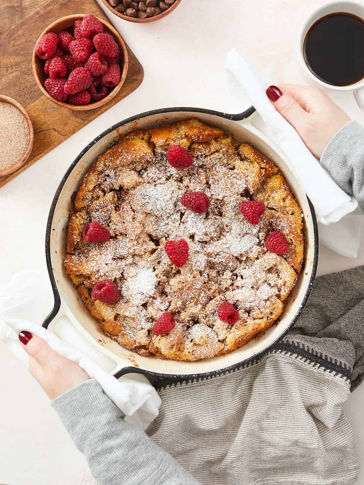 Image verticale d'un pudding au pain cuit dans un plat rond en fonte émaillée blanche, garni de sucre en poudre et de framboises fraîches. Il y a une planche à découper dans le coin supérieur gauche du cadre avec un bol en bois de framboises et à côté se trouve une tasse de café partiellement hors cadre.