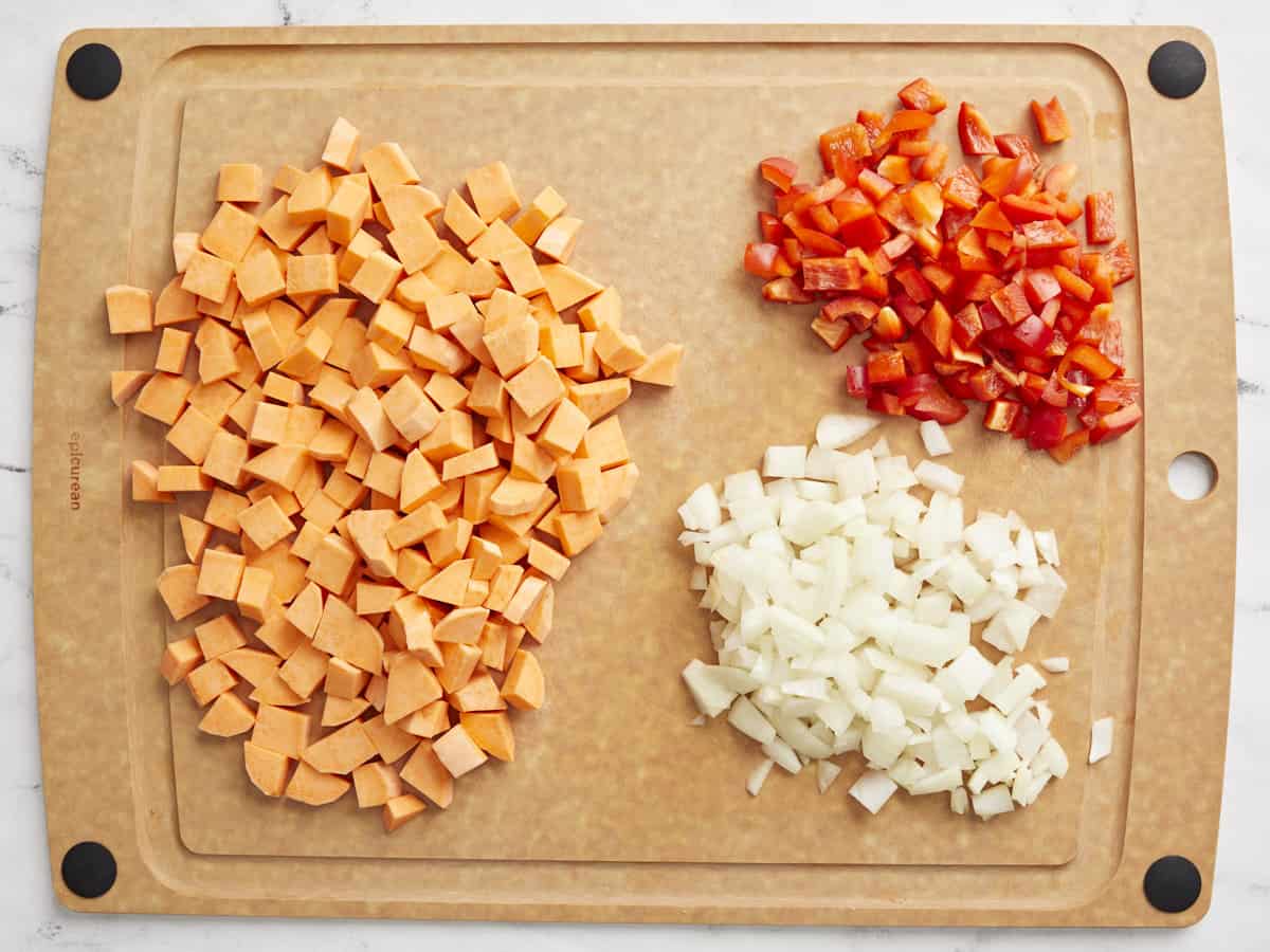 Patate douce coupée en dés, poivron rouge coupé en dés et oignon coupé en dés sur une planche à découper.