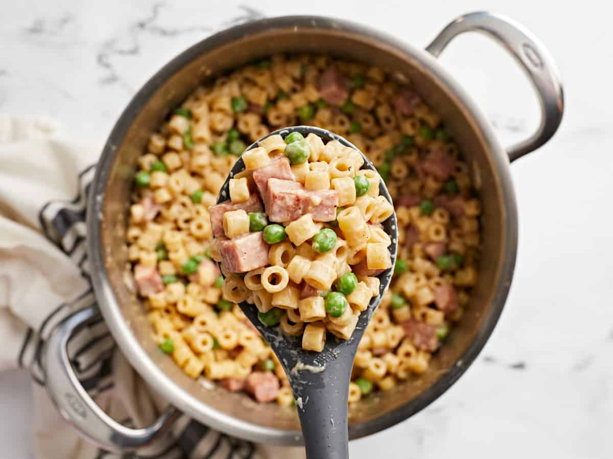 Gros plan de pâtes au jambon et aux petits pois dans une cuillère tenue au-dessus de la casserole.