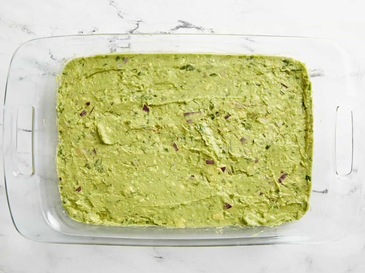 Le guacamole s'étale uniformément sur les haricots frits dans une cocotte en verre.