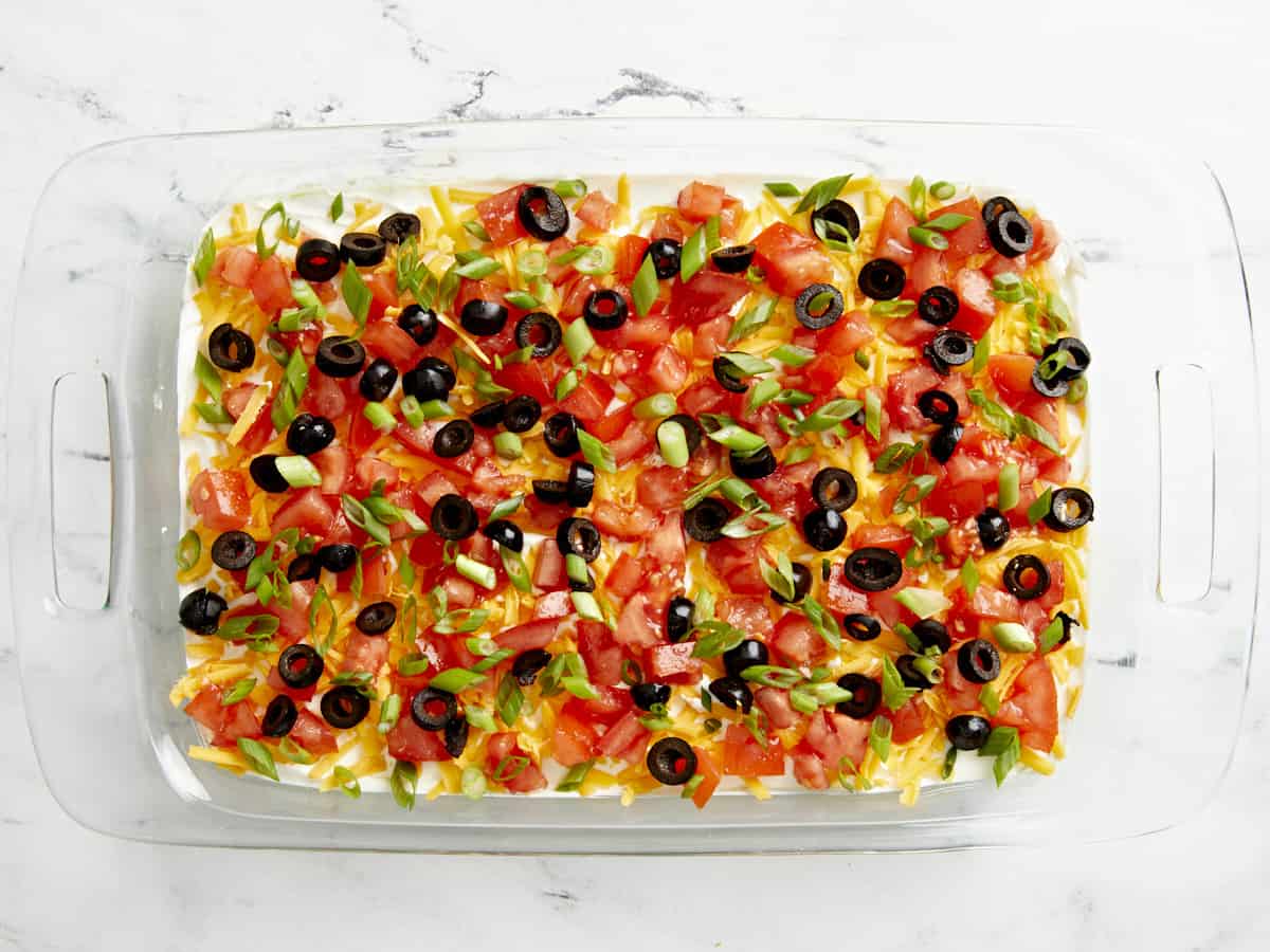 Tomates hachées, olives noires et oignons verts tranchés ajoutés et répartis uniformément sur la couche de fromage râpé.
