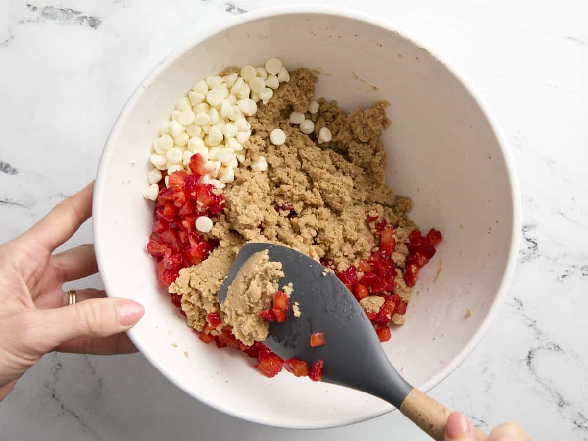 plier les morceaux de fraises et les pépites de chocolat blanc dans la pâte à biscuits dans un bol blanc avec une spatule en caoutchouc.