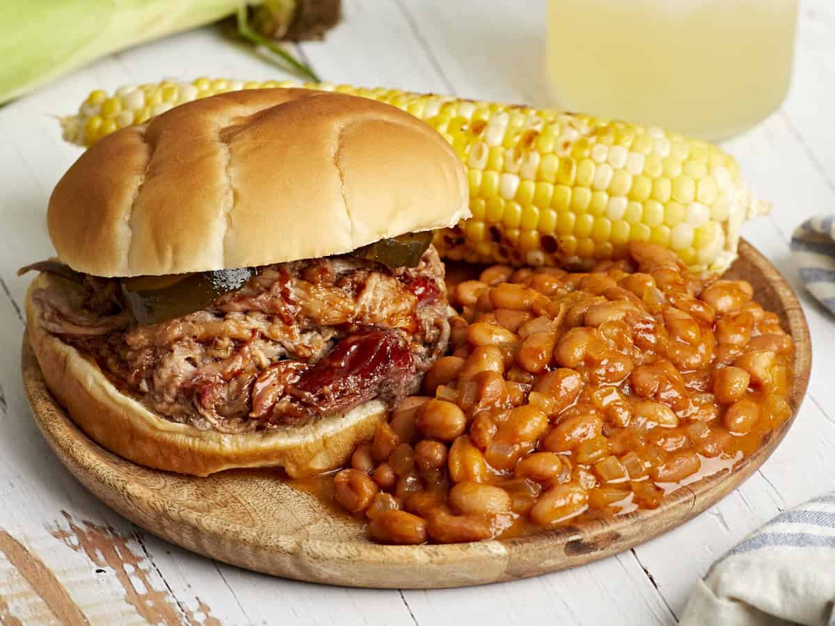 une portion de haricots cuits au four dans une assiette avec un sandwich au porc effiloché et des épis de maïs.