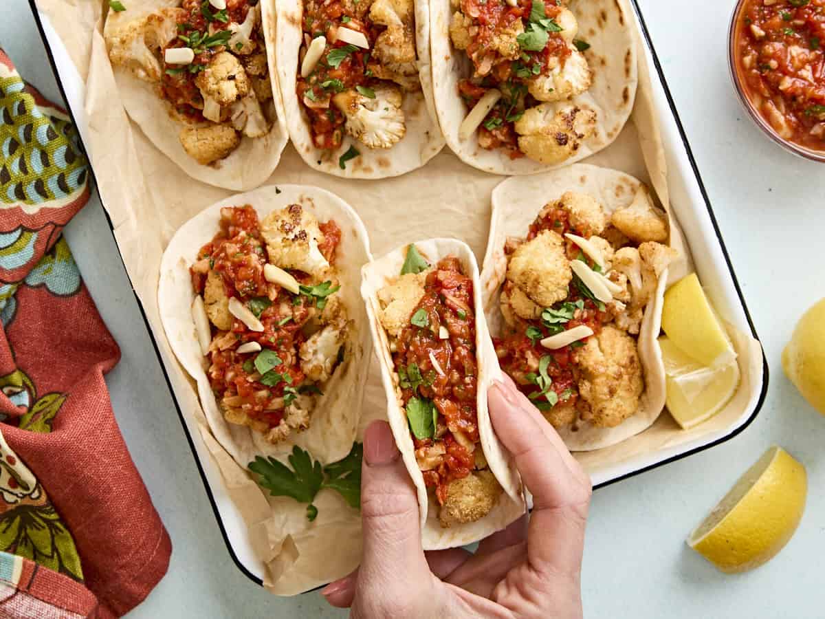 Une main prenant un taco dans un plat de cuisson rempli de tacos au chou-fleur avec de la sauce romesco
