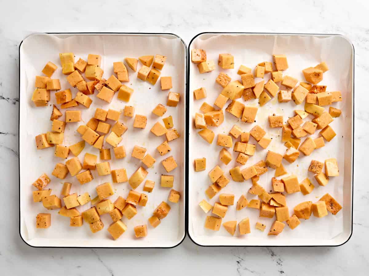 Cubes de courge musquée assaisonnés sur deux plaques à pâtisserie tapissées de papier sulfurisé.