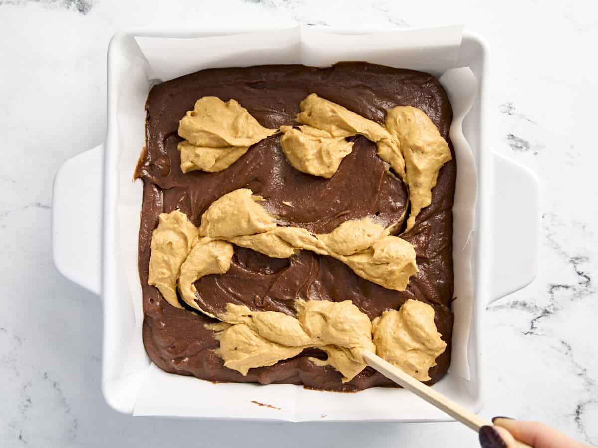 Un cure-dent étalant la garniture au fromage à la crème de citrouille sur la pâte à brownie dans un plat allant au four.