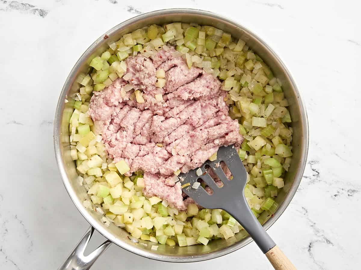 Saucisse hachée ajoutée aux légumes ramollis dans une poêle, brisée par une spatule.