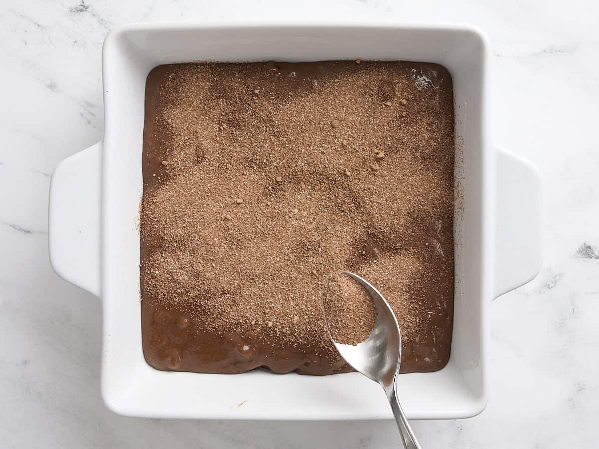 Une cuillère saupoudrant le mélange de sucre et de cacao sur la pâte à gâteau au pudding au chocolat dans un moule à gâteau.