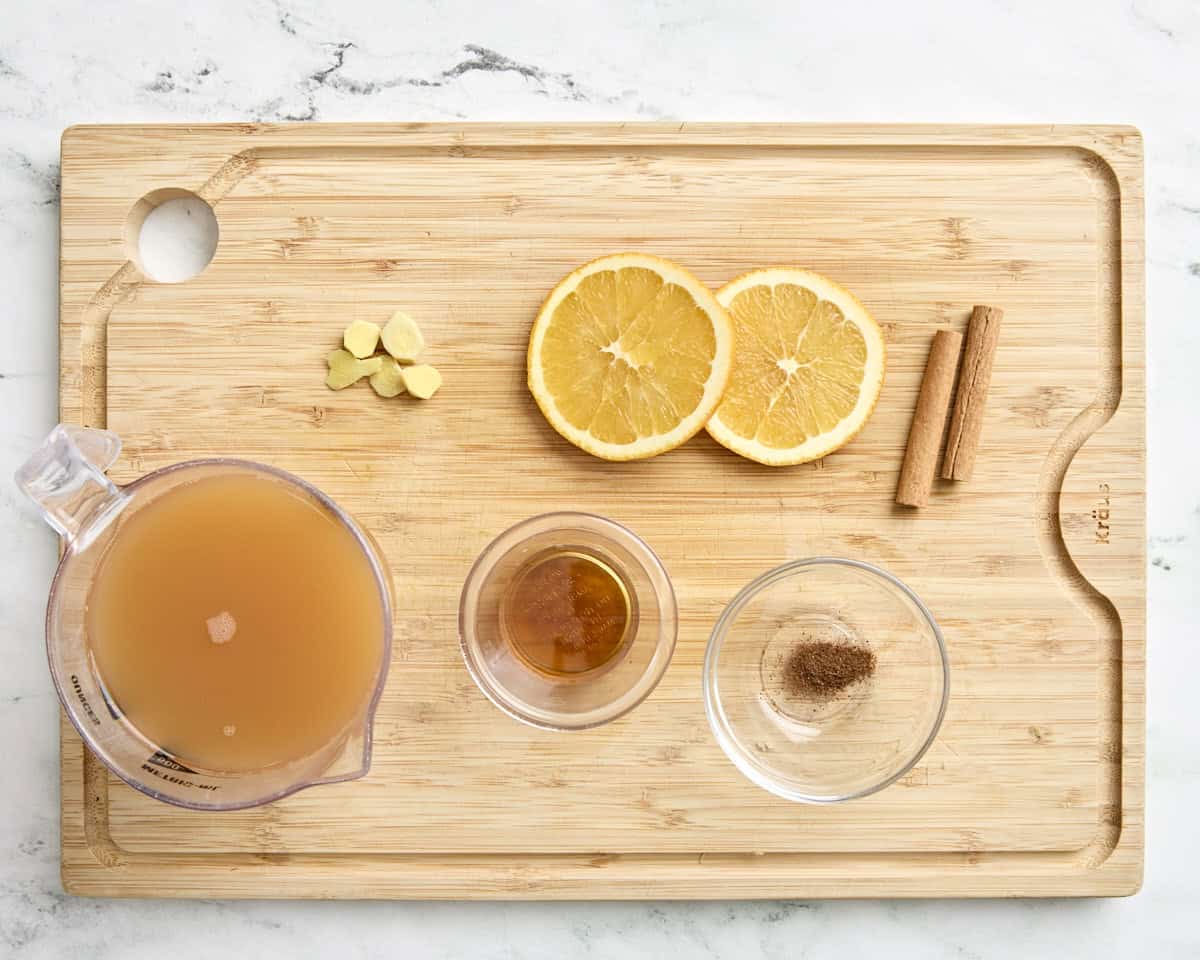 Cidre de pomme, tranches d'orange, gingembre frais haché, sirop d'érable, muscade et bâtons de cannelle sur une planche à découper en bois.