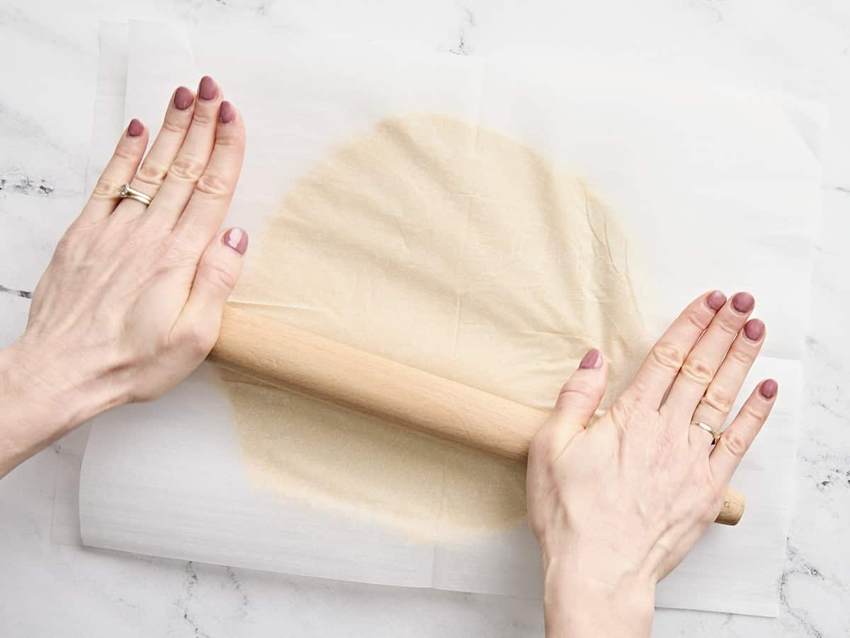 Mains étalant la pâte à biscuits au sucre entre deux feuilles de papier sulfurisé à l'aide d'un rouleau à pâtisserie.
