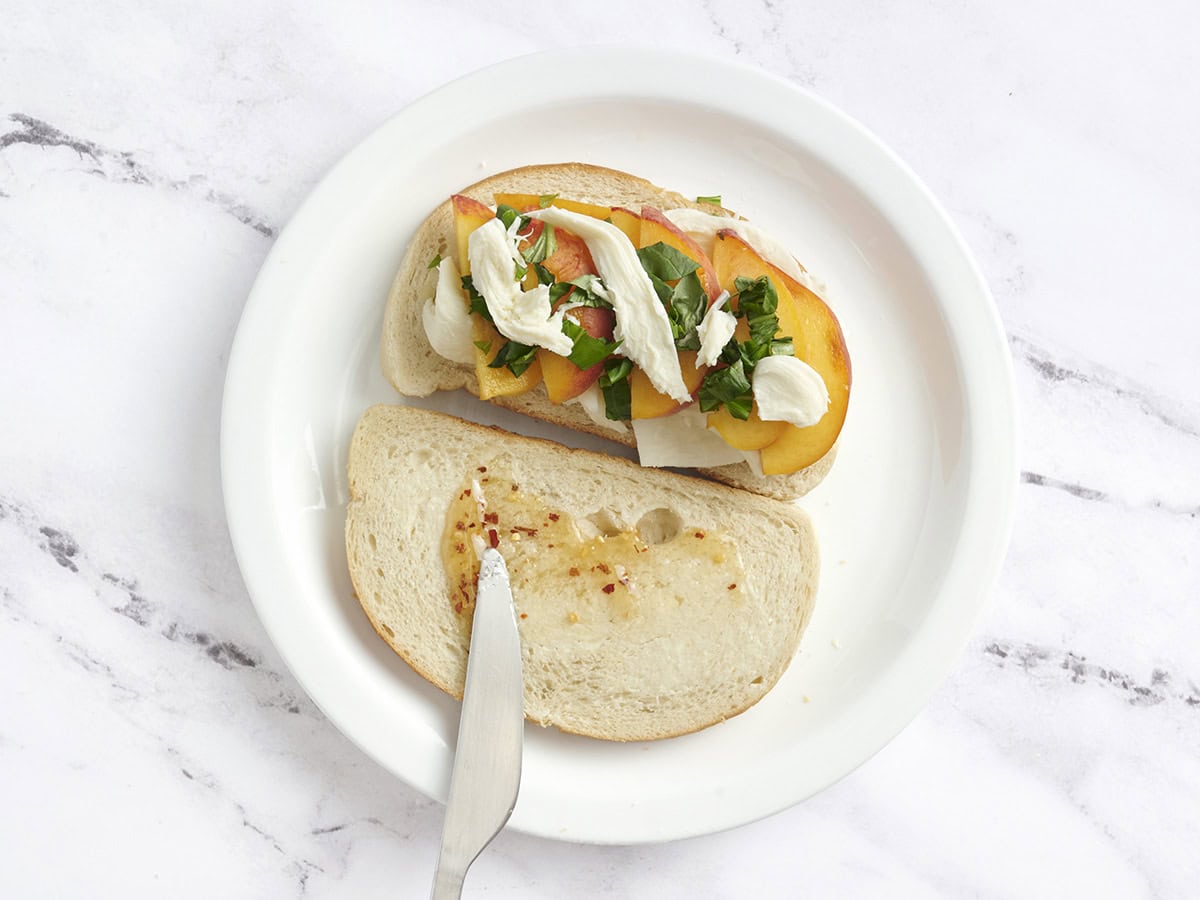 Deux tranches de levain beurré sur une assiette, avec l'un garni de pêches, de mozzarella et de basilic et l'autre a du miel étalé sur le dessus.