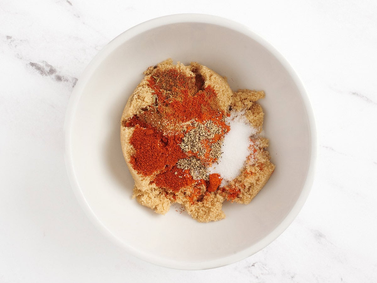 Sucre brun, cayenne, poudre d'ail, paprika, sel, poivre noir, gousses, cannelle et thym dans un bol à mélanger.