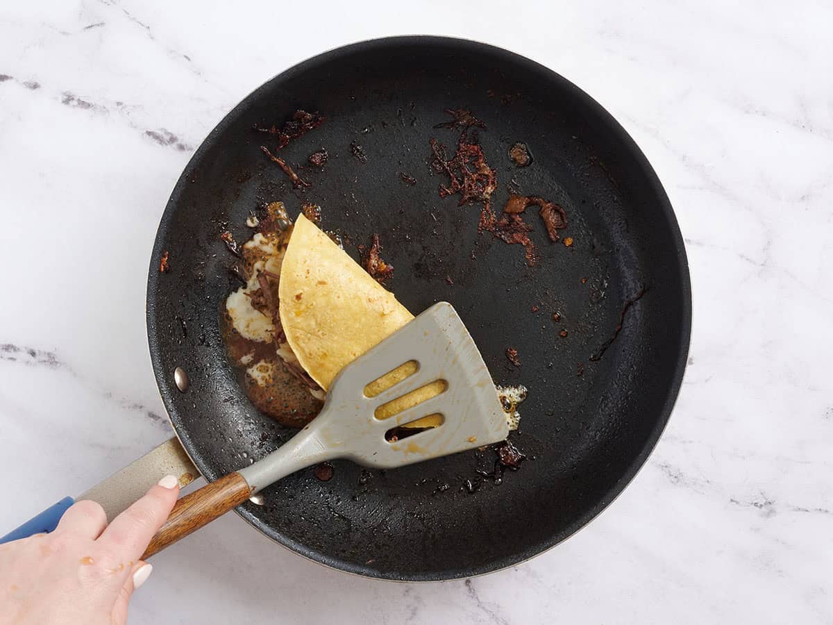 Une spatule appuyant sur un taco Birria dans une poêle.
