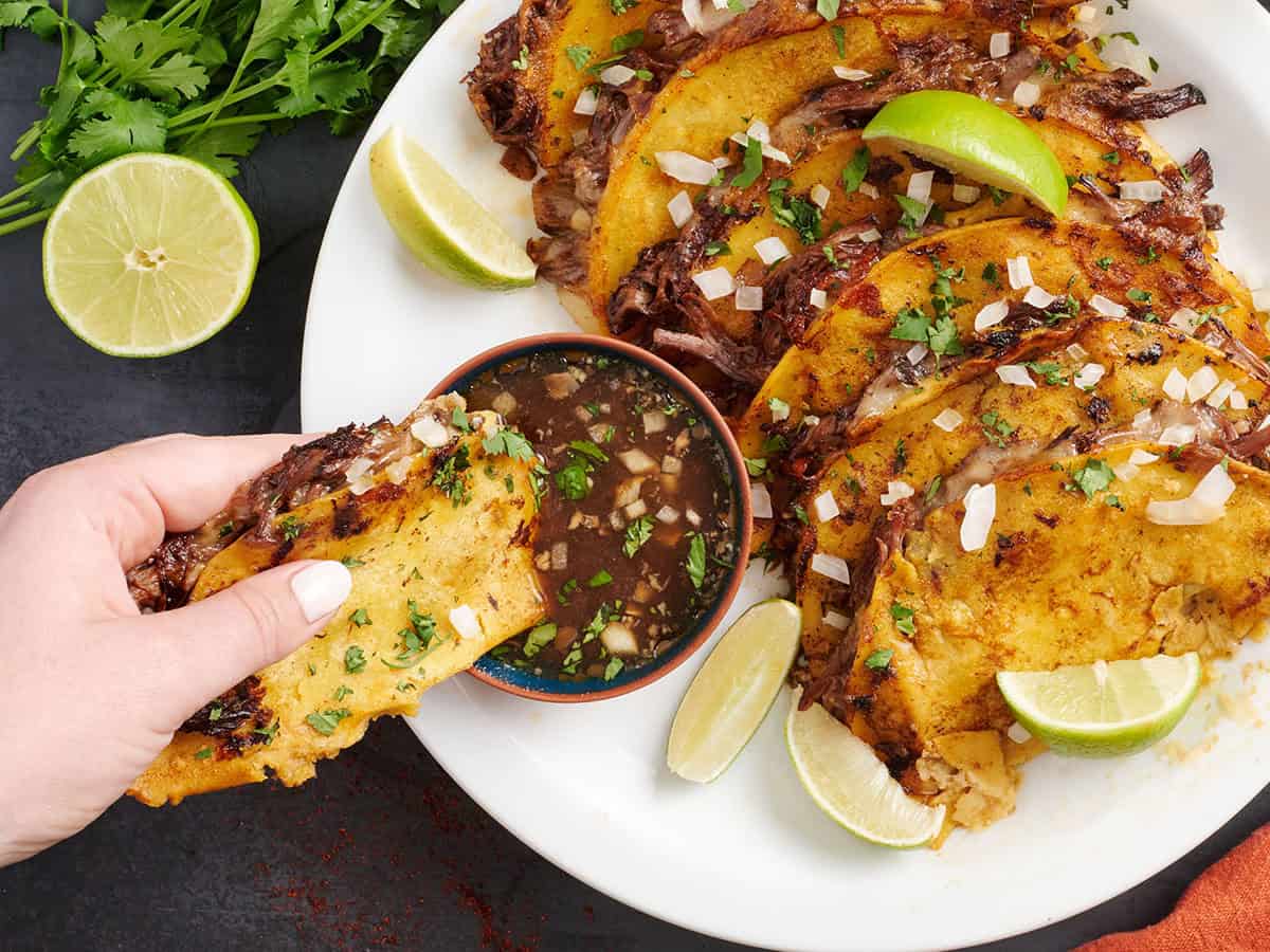 Vue aérienne des tacos de Birria dans une assiette avec une main en trempant dans un pot de consomme de bœuf.
