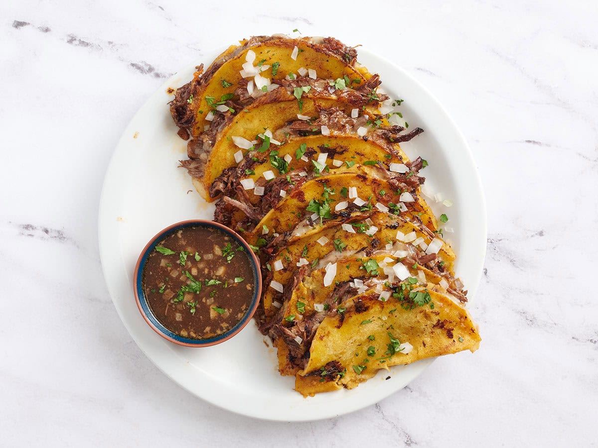 Un plateau de tacos de birria faits maison avec un pot de bouillon de bœuf pour tremper.