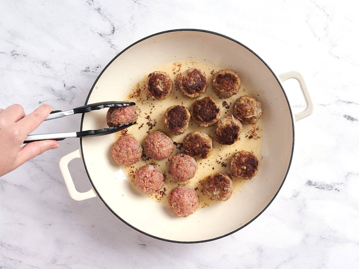 Boulettes de viande suédoises cuisant dans une casserole, avec des pinces les retournant.