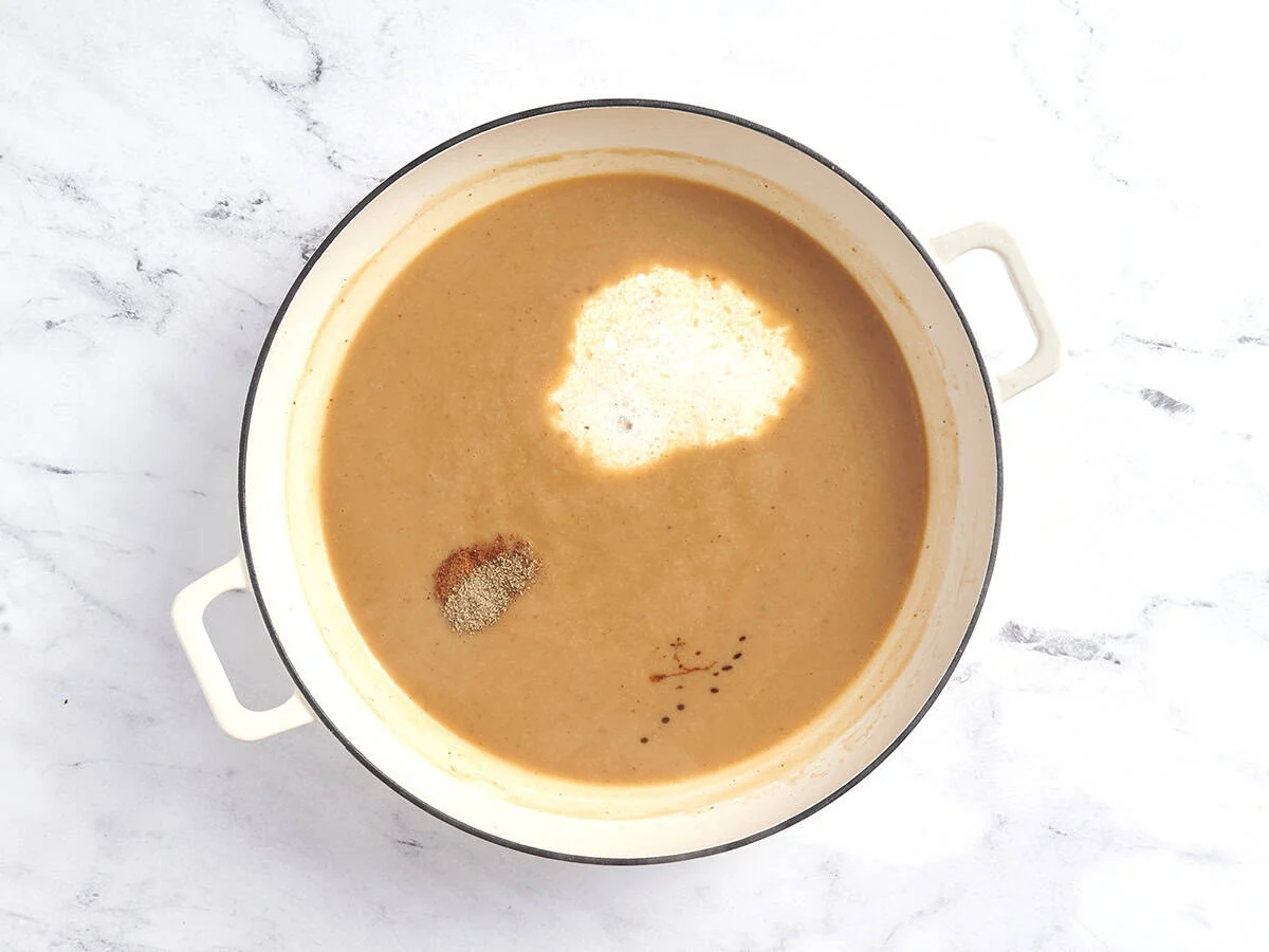 Épices et crème ajoutées à un roux dans une casserole pour obtenir une sauce crémeuse.