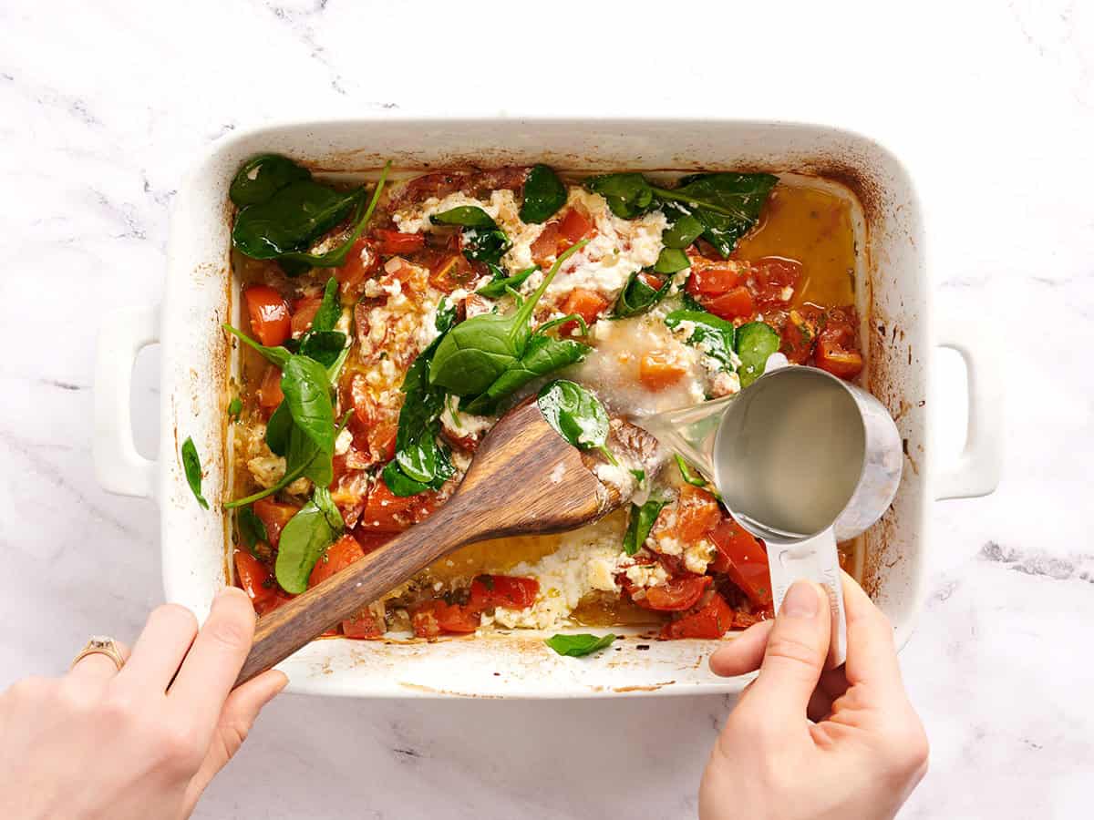 Une main versant l'eau réservée aux pâtes dans un plat allant au four composé de tomates cuites au four et de fromage feta.