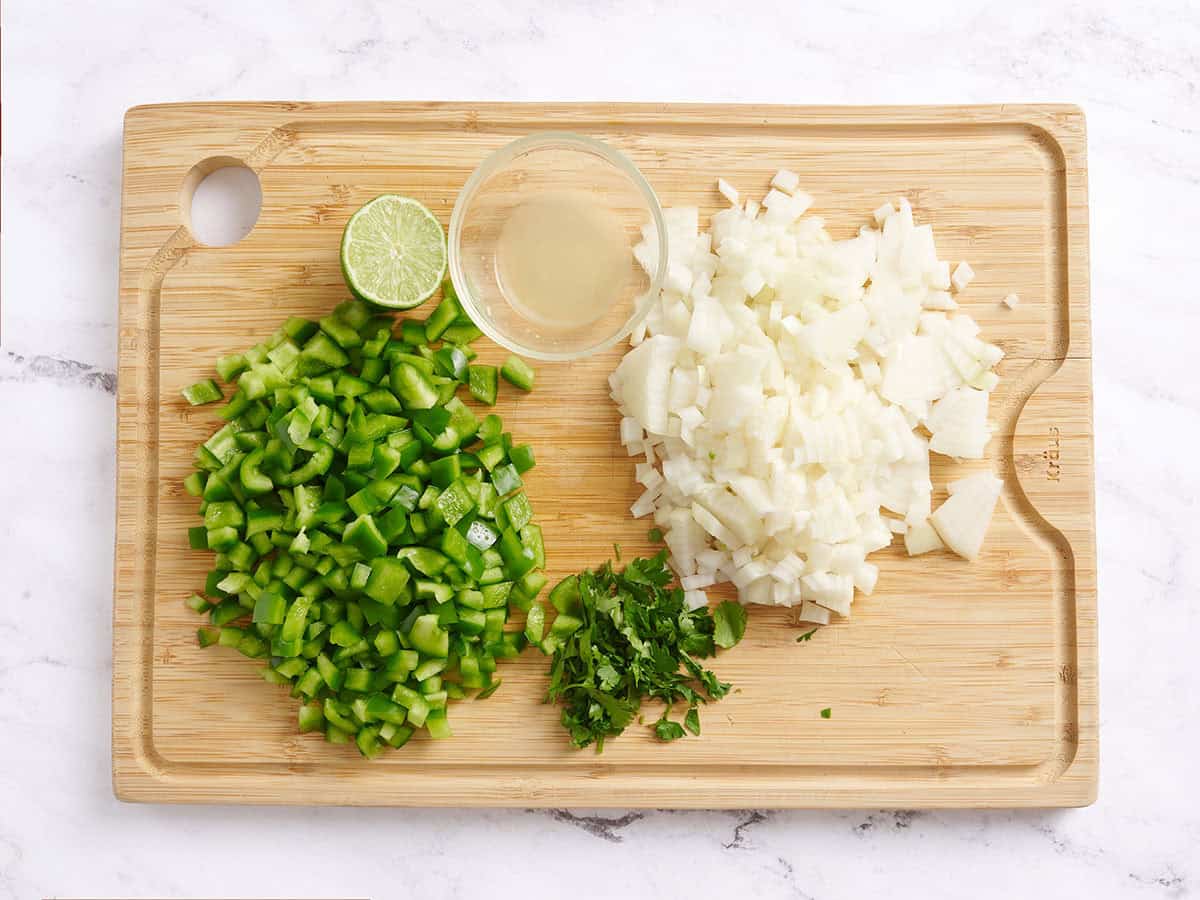 Poivron vert coupé en dés, oignon coupé en dés, coriandre hachée et citron vert sur une planche à découper en bois.