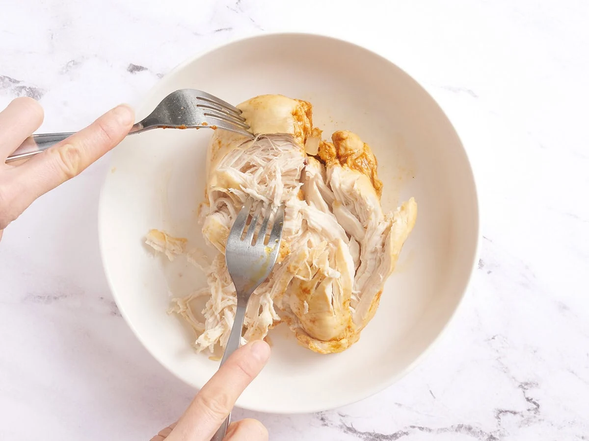 Mains utilisant deux fourchettes pour déchiqueter les poitrines de poulet cuites dans un bol.