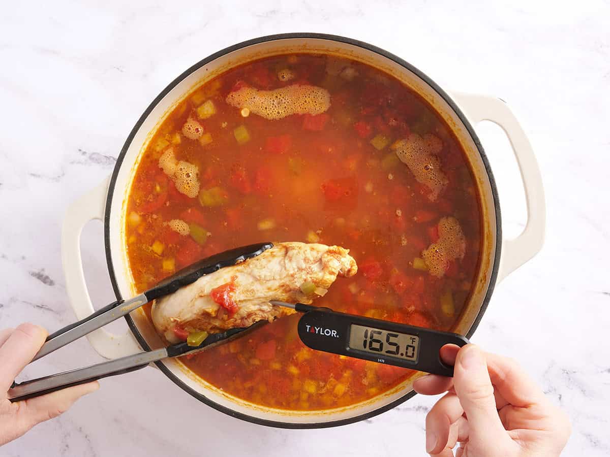 Pinces tenant une poitrine de poulet cuite sur une casserole de soupe taco au poulet et un thermomètre à viande vérifiant la température interne du poulet.