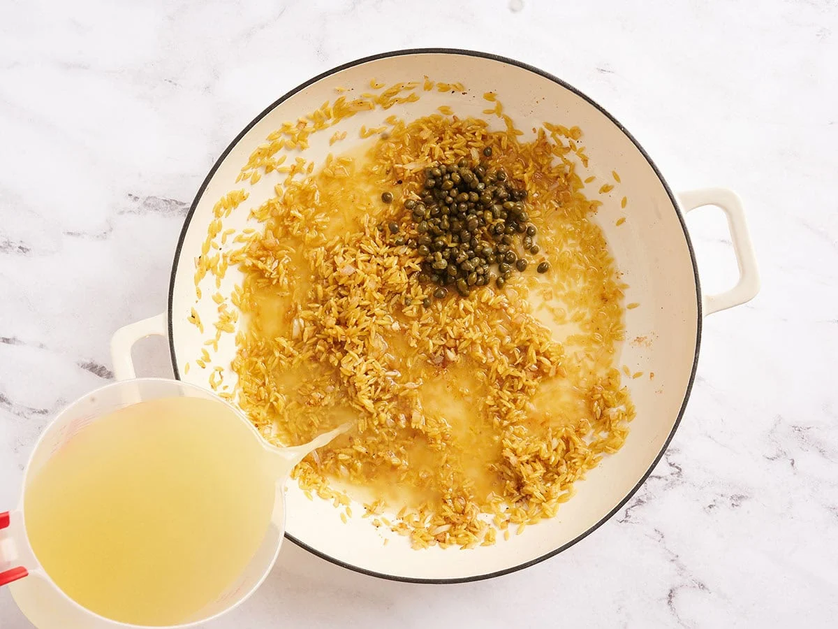 Câpres et bouillon ajoutés dans une poêle avec de l'orzo.