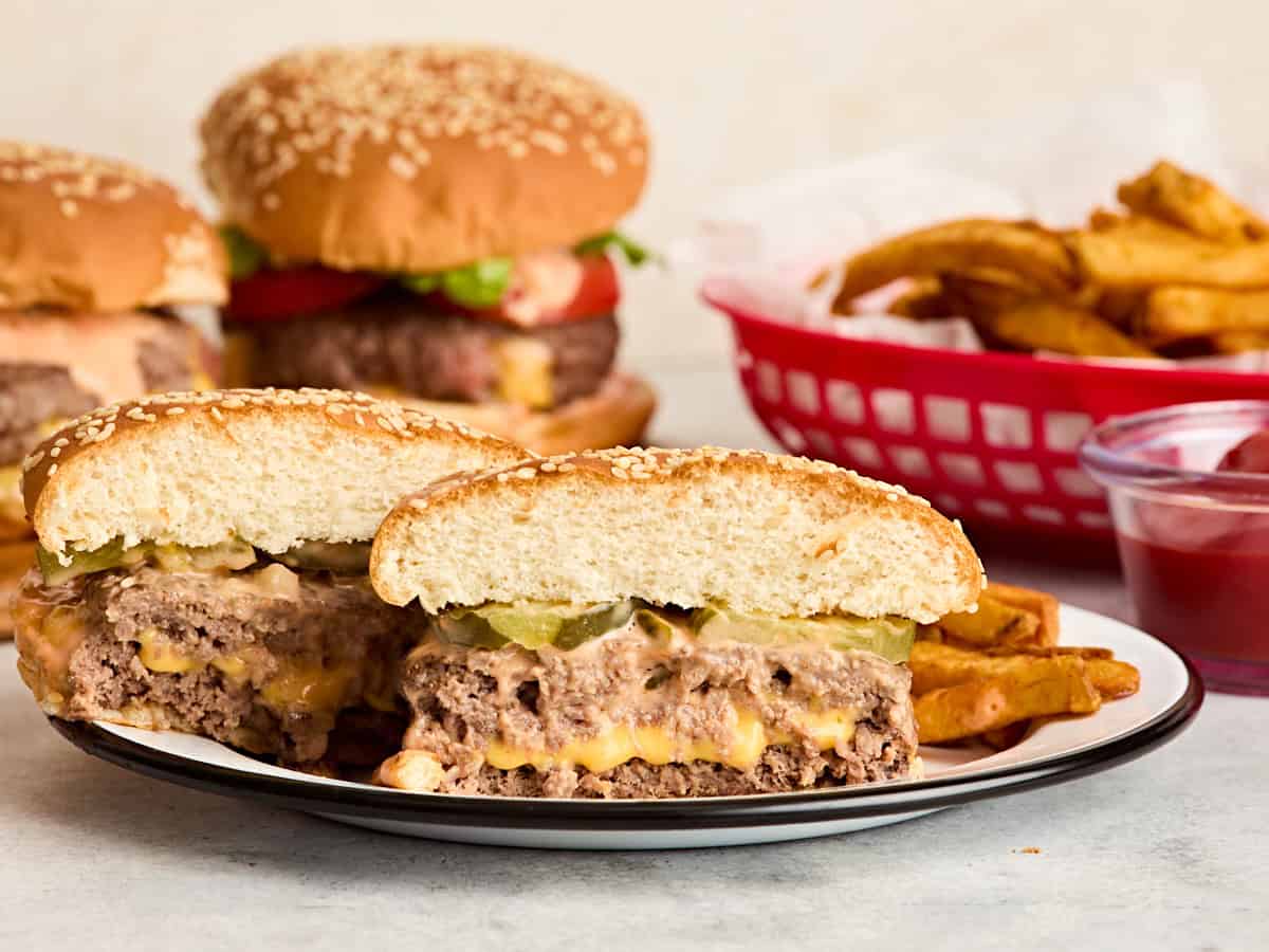 Vue latérale d'un hamburger lucy juteux coupé en deux sur une assiette avec plus de hamburgers et un panier de frites en arrière-plan.
