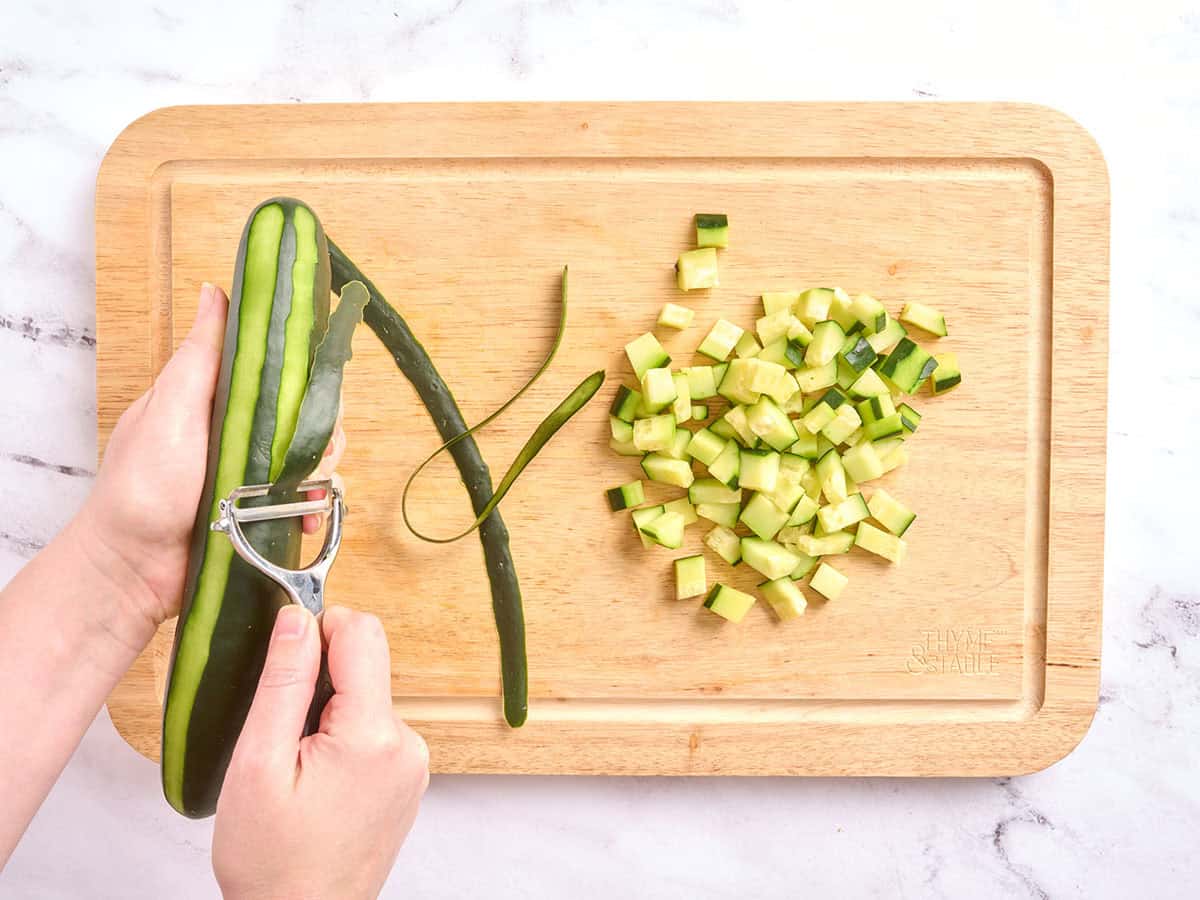 Mains épluchant un concombre, à côté d'une planche à découper en bois avec des dés de concombre dessus.