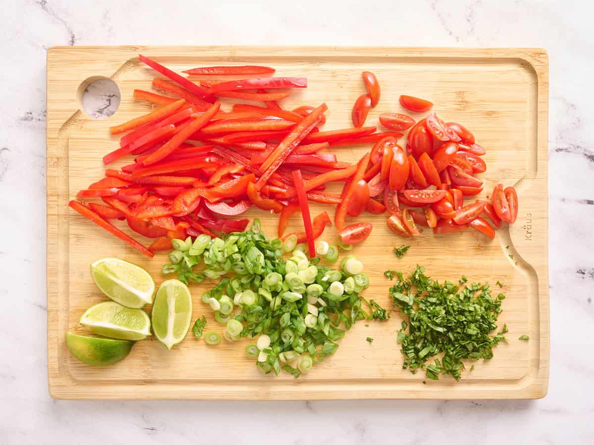 Poivron rouge tranché, oignon vert tranché, quartiers de lime en quartiers, tomates raisins tranchées et basilic émincé sur une planche à découper en bois.