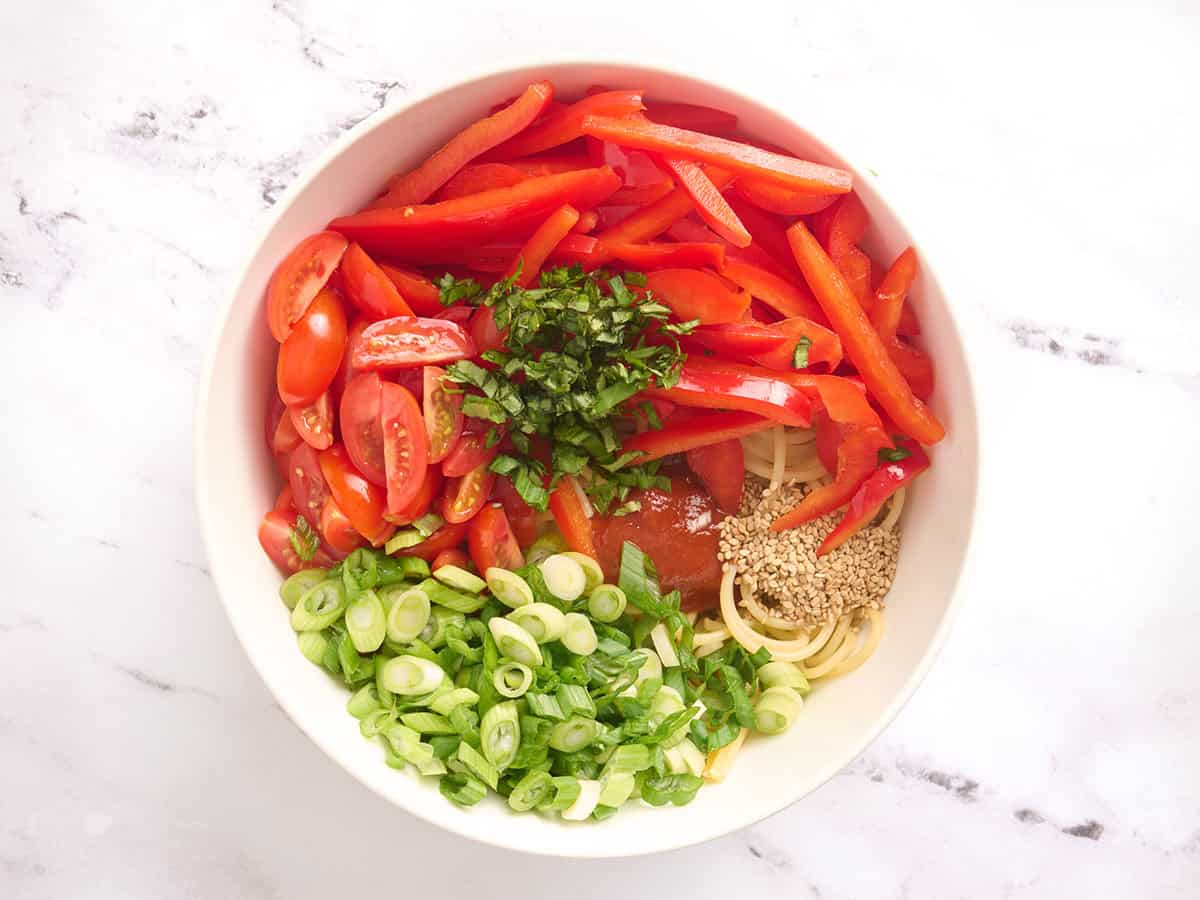 Poivron rouge tranché, oignons verts tranchés, basilic émincé, tomates raisins tranchées et spaghettis dans un bol.
