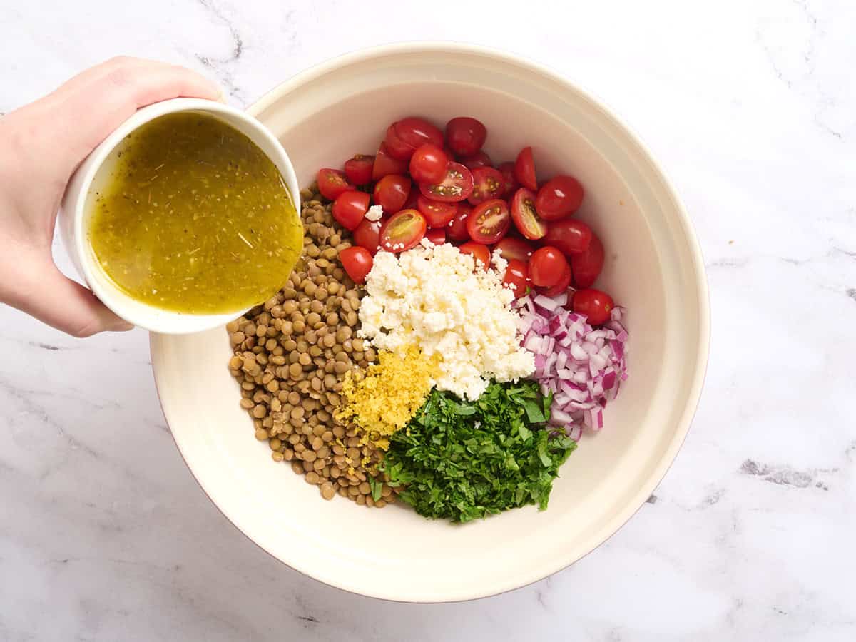 Vinaigrette versée dans un bol avec tomates, feta, persil, oignon rouge et lentilles cuites.