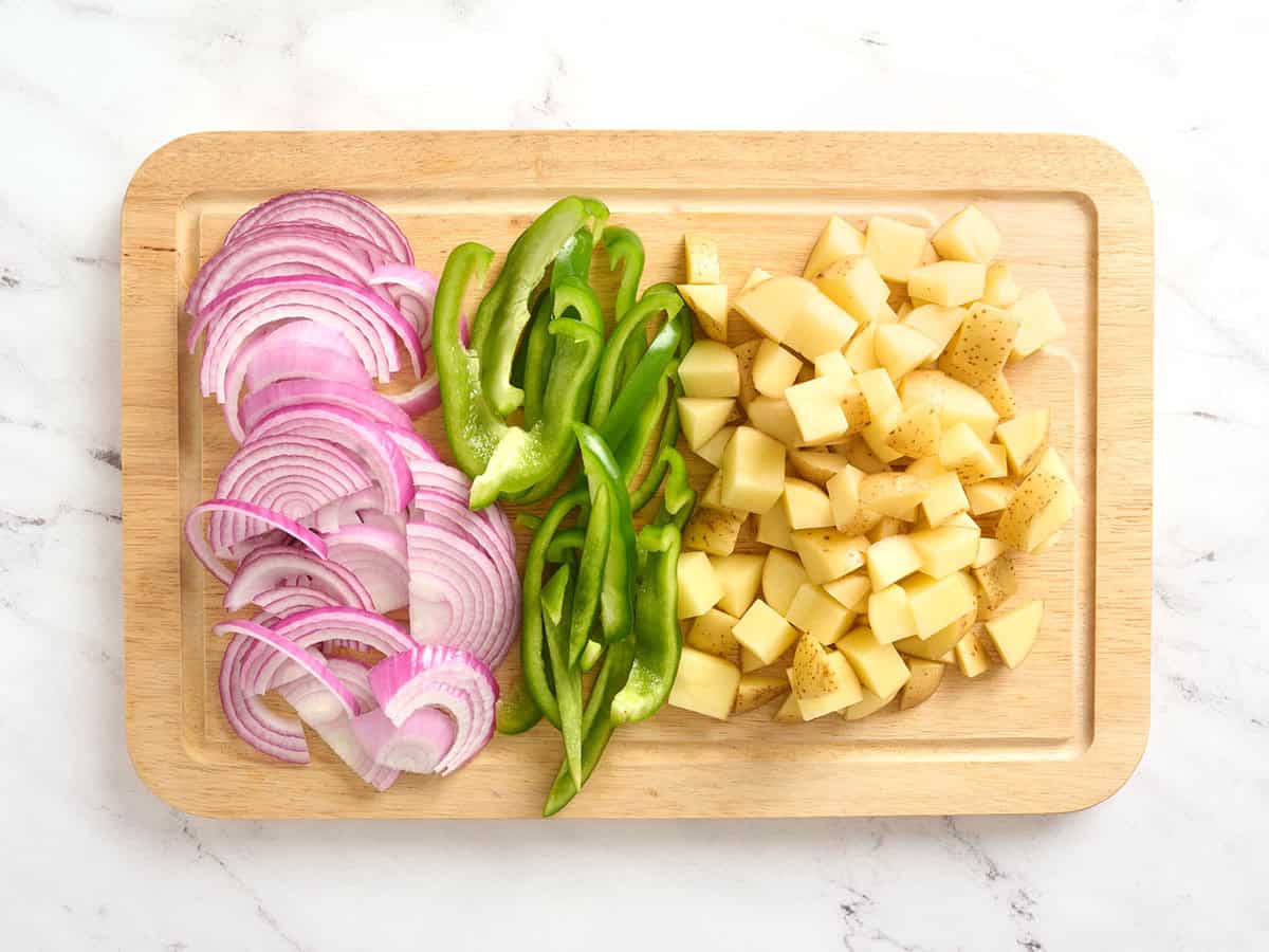 Oignons rouges tranchés, poivrons verts et pommes de terre en dés sur une planche à découper.