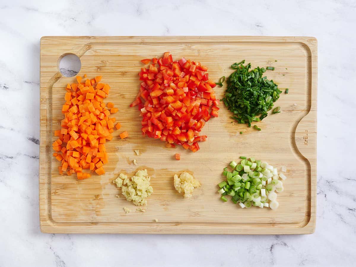 Carottes coupées en dés, poivrons, oignons verts, ail et gingembre émincés sur une planche à découper en bois.